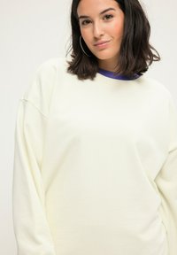 Sudadera de color crema claro con corte holgado, textura suave y mangas anchas; superpuesta sobre una camiseta morada en el cuello.