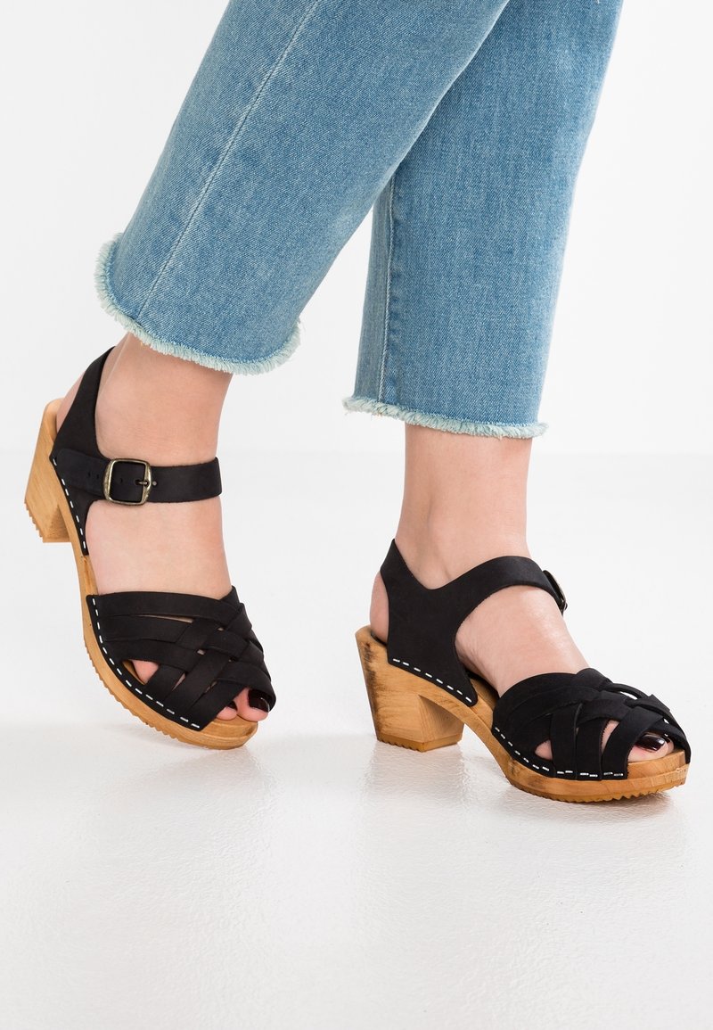 Pieds chaussés de sandales noires à talons en bois et à brides, portées avec un jean bleu clair à ourlet effiloché et court, sur un sol blanc.