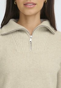 Beige strikket genser med ribbestruktur, med glidelås i halsen og stor krage. Sleek sølvglidelås fremhever designet.
