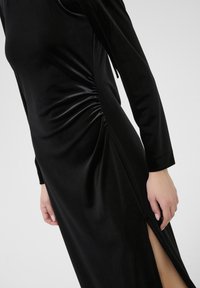 Robe en velours noire à manches longues, avec des fronces sur le côté et une fente haute à la cuisse. La texture lisse et le design ajusté mettent en valeur sa silhouette.