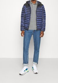 Blå puffjacka med svart huva, grå polo och ljusblå jeans. Kombinerad med vita sneakers med gröna och blå accenter.