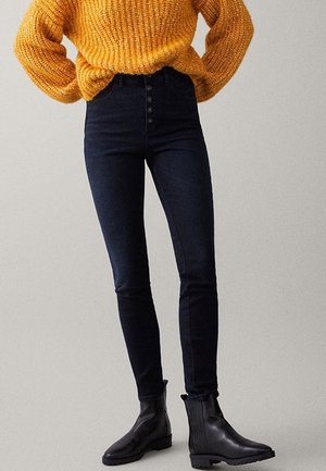Personne portant un jean skinny noir taille haute avec braguette boutonnée, des bottines noires et un pull en grosse maille jaune moutarde.