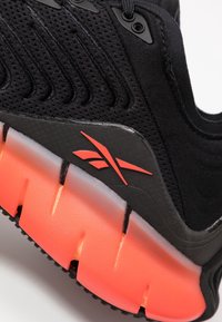 Chaussure de sport noire avec une tige en maille respirante, un design rainuré distinctif et une semelle amortie avec des accents orange prononcés.