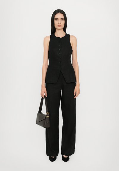 Black linen sleeveless top with a front button closure, paired with loose-fitting black trousers, and a dark handbag. Smooth texture, minimal design.