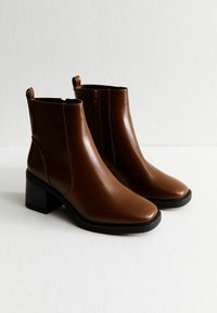 Bottes chelsea en cuir marron avec une finition lisse, bout carré et talon bloc. Présente une fermeture éclair sur le côté et un détail de couture subtil.