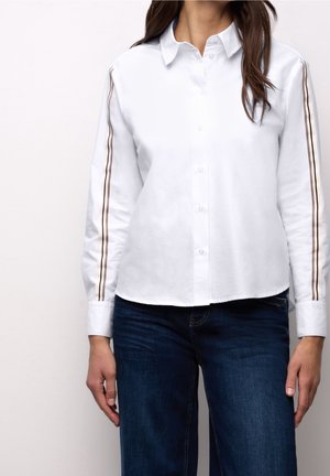 Femme portant une chemise blanche boutonnée avec de fines doubles rayures sur les manches, associée à un jean bleu foncé, debout devant un fond uni.