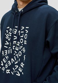 Persona con una sudadera azul marino con texto distorsionado en blanco, dispuesto en un patrón circular en la parte frontal y cordones colgando.