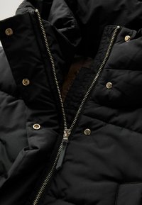 Gros plan sur une doudoune noire, présentant un col haut, une fermeture éclair dorée et des boutons-pression, avec un revêtement doux visible à l'intérieur.