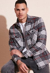 Cămașă din flanel în carouri, cu culorile negru, gri și roșu, cu închidere cu nasturi, două buzunare la piept și un croi lejer. Pur și simplu cu un tricou alb dedesubt.