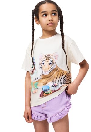 Jente med flettehår iført en kremfarget t-skjorte med et tigerguttmotiv og pastell-lilla volangshorts, som ser til siden.