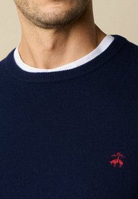 Granatowy sweter z dzianiny z okrągłym białym kołnierzykiem, z małym czerwonym haftem przedstawiającym dwa zwierzęta. Miękka tekstura, casualowy styl.