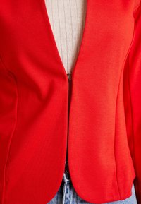 Blazer rouge en tissu lisse, avec un col accentué, une fermeture éclair et une coupe ajustée, associé à un haut beige texturé et un jean en denim.