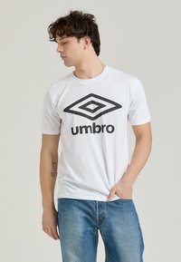 La t-shirt bianca in cotone presenta un grande logo Umbro nero sul davanti. Ha maniche corte e un design a girocollo con una texture liscia.