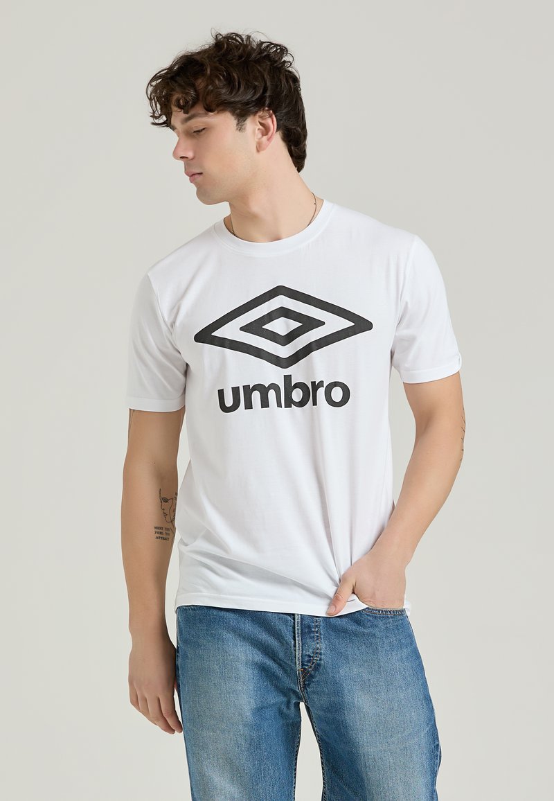 La t-shirt bianca in cotone presenta un grande logo Umbro nero sul davanti. Ha maniche corte e un design a girocollo con una texture liscia.