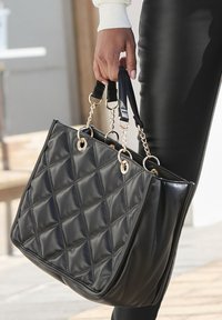 Schwarze, gesteppten Tragetasche aus glänzendem Material mit goldenen Akzenten und Kettenverzierungen, ausgestattet mit stabilen Schulterriemen und einem geräumigen Inneren.