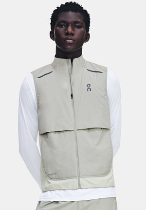 Jeune homme portant un gilet sans manches gris clair zippé par-dessus une chemise blanche à manches longues, debout devant un fond uni.