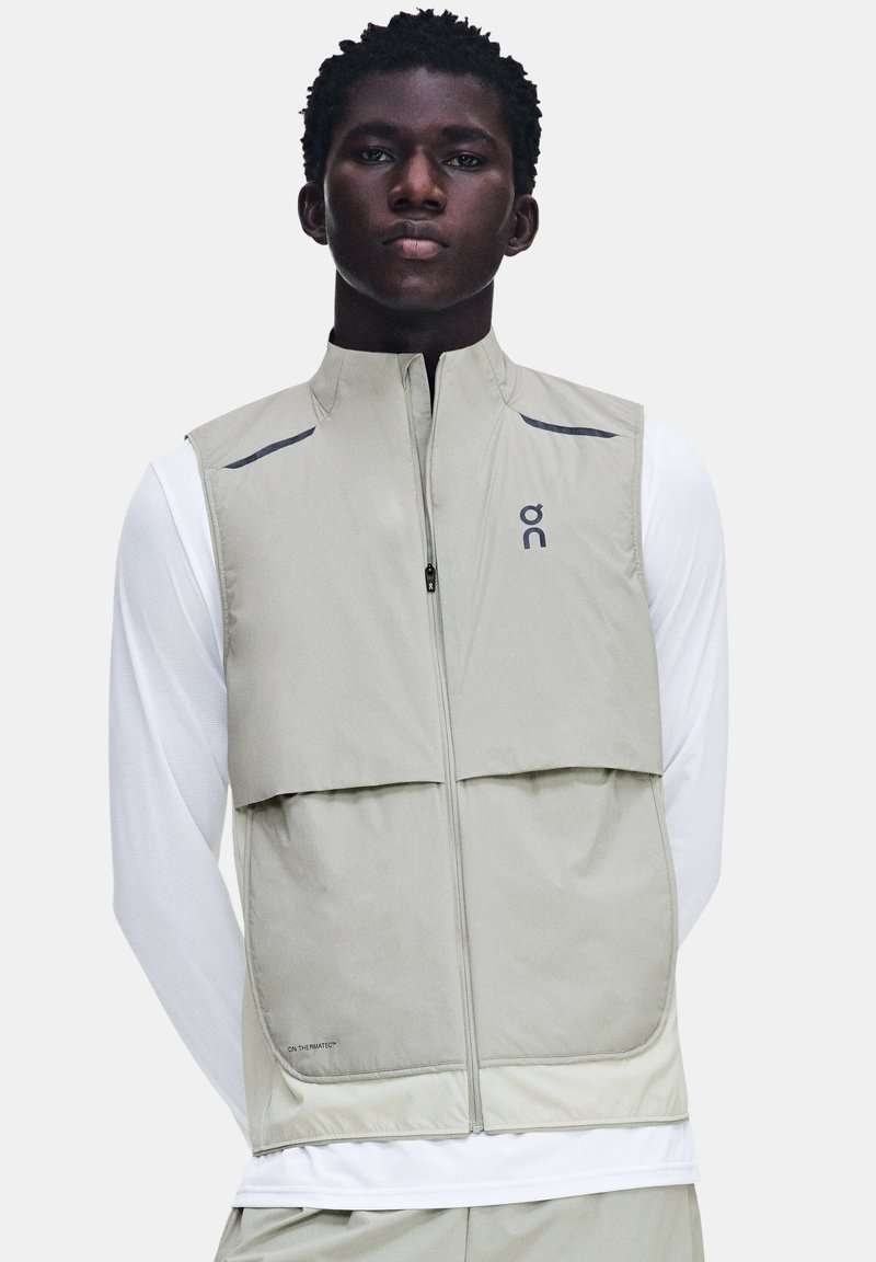Jeune homme portant un gilet sans manches gris clair zippé par-dessus une chemise blanche à manches longues, debout devant un fond uni.