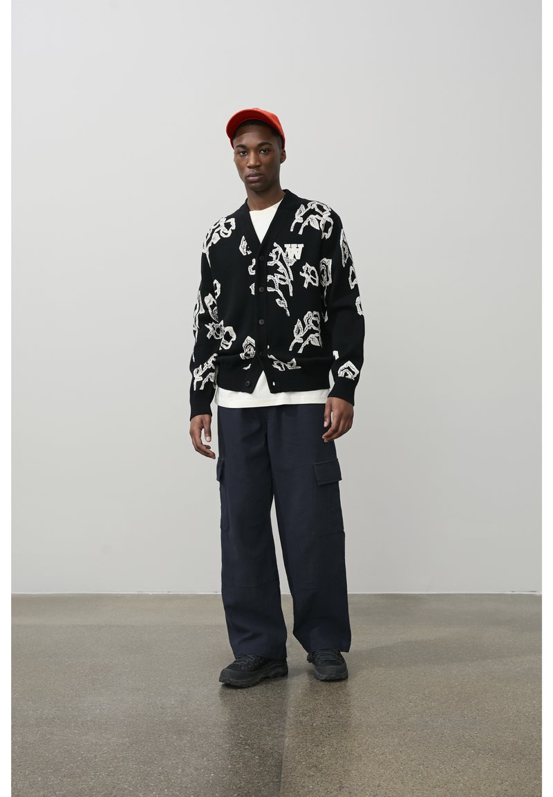 Čovjek u crnom kardiganu s bijelim apstraktnim uzorcima, bijelom košuljom, tamnoplavim cargo hlačama, crnim cipelama i crvenom kapa, stoji na uglačanoj podlozi.