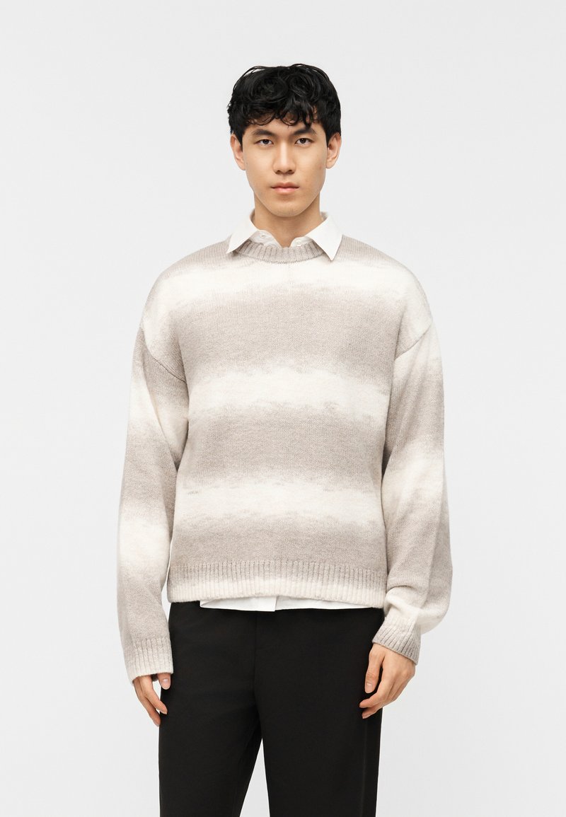 Homme portant un pull beige et blanc à rayures sur une chemise blanche à col, avec un pantalon noir, se tenant devant un fond clair uni.