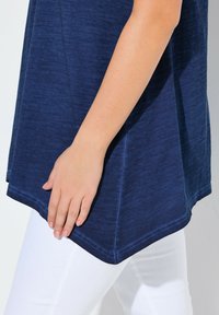 Tummansininen paita, jossa on tekstuuri ja kaareva helma, yhdistettynä valkoisiin housuihin. Käsi lepää kevyesti kankaalla.