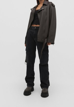 Frau trägt dunkelbraune Lederjacke über schwarzem Crop-Top, schwarze Cargohose und klobige schwarze Stiefel vor schlichtem grauem Hintergrund.