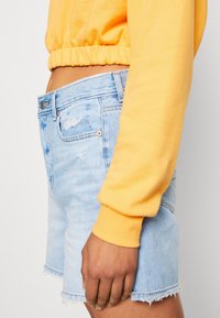 Shorts in denim azzurro chiaro con orli sfrangiati e zone distressate. Abbinati a una felpa gialla con vita elasticizzata. Vista ravvicinata di lato.