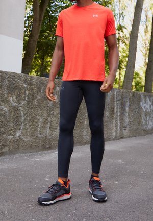 Mann trägt ein hellorangefarbenes kurzärmeliges Hemd, schwarze Leggings und schwarz-orangefarbene Laufschuhe, steht im Freien auf dem Bürgersteig.