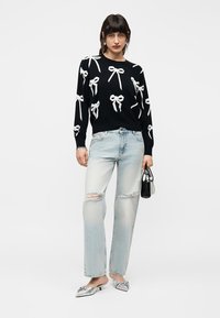 Femme portant un pull noir orné de nœuds blancs, un jean clair déchiré, des chaussures argentées à bout pointu, et tenant un petit sac à main noir.