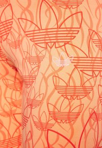 Orange tyg med ett upprepat mönster av konturerade klöverformer i mörkare orange, med en vit klöverlogotyp i det nedre högra hörnet.