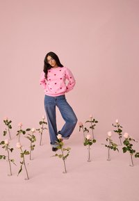 Žena v ružovom svetri so vzorom srdiečok a džínsach stojí bosá medzi ružovými ružami vo sklenených váza proti bledo ružovému pozadiu.