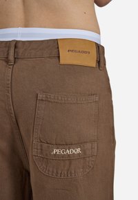 Smeđe pamučne hlače imaju stražnji džep s izvezenim logotipom i kožnu oznaku na struku s imenom brenda.