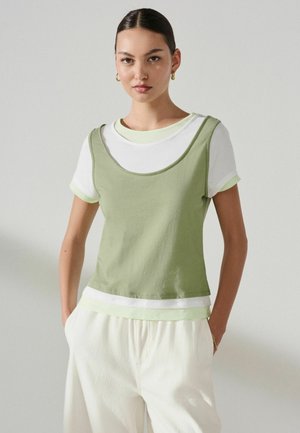 Young woman with sleek hair wearing gold hoop earrings, a white t-shirt layered with a light green sleeveless top, and loose white pants.