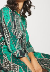 Grön mönstrad klänning med trekvartslånga ärmar, prydd med paisley- och geometriska mönster i kontrasterande färger. Mjuk, slät textur.