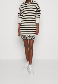 Personne portant un sweat à capuche rayé noir et blanc avec une jupe à motifs noir et blanc et des baskets blanches, sur un fond uni.