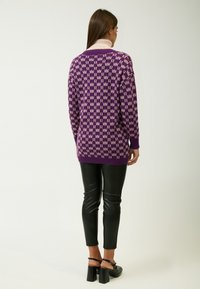 Lila Pullover mit einem pinkfarbenen Muster, langen Ärmeln und lockerer Passform, kombiniert mit schwarzen Lederhosen und schwarzen Sandalen mit Absatz.