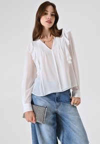Camicia bianca trasparente con maniche arricciate, scollo a V e pieghe frontali, abbinata a jeans di denim blu chiaro a vita alta e una borsa a tracolla testurizzata.