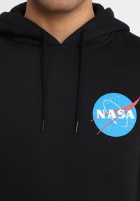 Sudadera negra con cordones. Presenta un logo azul de la NASA con texto blanco y un acento rojo. Tejido suave, corte estándar.