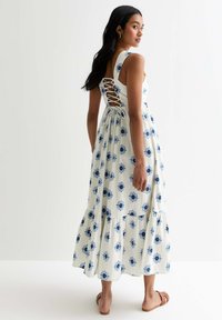Blommig midi-klänning i vitt tyg med blå blommönster, med volangkjol och snörning bak. Stylist som bärs med platta sandaler.