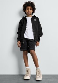 Felpa nera con zip e logo sul petto, sotto una t-shirt bianca, abbinata a pantaloncini neri e scarpe alte beige.