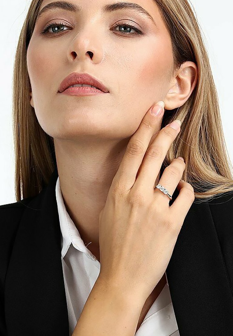 Silberner Ring mit strukturiertem Band und kleinen Edelsteinen, getragen an einer Hand, die den Hals berührt. Modell gekleidet in einem schwarzen Blazer und einem weißen Hemd.