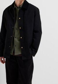 Musta puuvillatakki, jossa on kullanväriset nappikoristeet ja samettikaulus, päällä vihreä paita ja mustat housut. Yksinkertainen malli ja rento istuvuus.