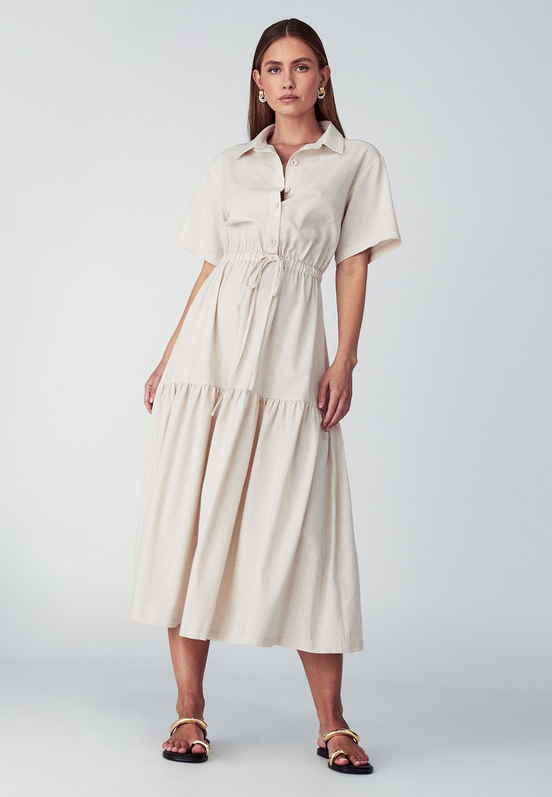 Šviesiai rudas, trumpomis rankovėmis, mygtukais užsegamas suknelė su elastinga juosta ir pakopiniu sijonu; glotnus paviršius, lengva medžiaga ir laisvas siluetas.