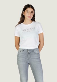 Jeune femme aux longs cheveux bruns portant un t-shirt blanc Armani Exchange et un jean gris clair, se tenant debout les mains derrière le dos.
