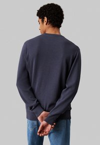 Maglione crewneck blu scuro con rifinitura testurizzata, maniche lunghe e polsini a coste, indossato con jeans blu. Visto dalla parte posteriore.