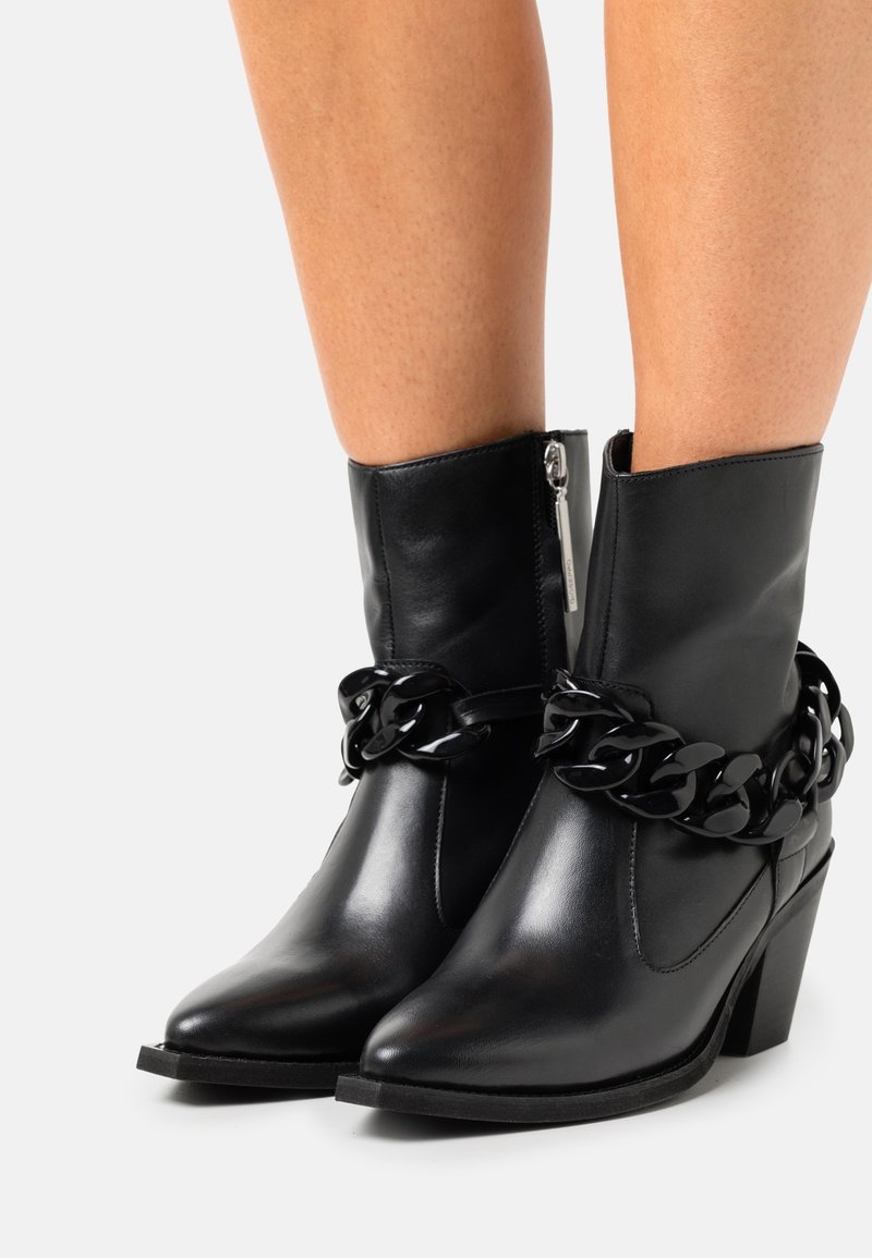 Bottines en cuir noir avec un gros détail chaîne, bouts pointus, fermeture éclair latérale et talons empilés portés sur jambes nues.