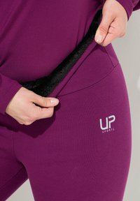 Händer som drar isär lila sportleggings för att avslöja svart fleecefoder inuti, som visar värme och textur, med "UP Sports"-logotypen.