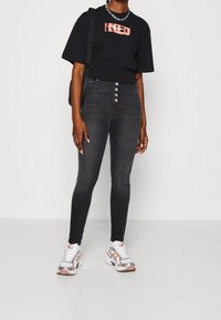 Camiseta gráfica negra, jeans ajustados de cintura alta en gris oscuro con cinco botones, y zapatillas deportivas multicolores con detalles en plateado.
