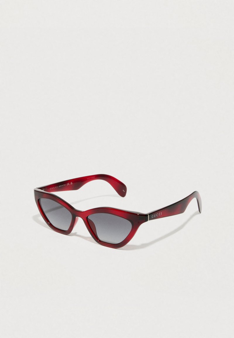 Ochelari de soare roșii, în formă de ochi de pisică, cu lentile de gradient întunecat. Fabricati din plastic neted, având brațe subțiri cu logo-ul "GUCCI" pe o parte.