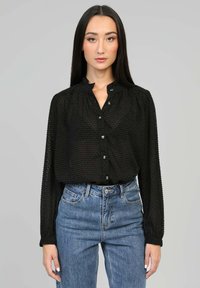 Blouse noire texturée avec un col froncé, une fermeture à boutons sur le devant et des manches longues ; portée avec un jean en denim bleu à taille haute.