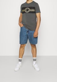 Šedé tričko s černým a zlatým pruhovaným logem, doplněné modrými denimovými šortkami. Oblečení zakončeno bílými teniskami a ponožkami s černými pruhy.